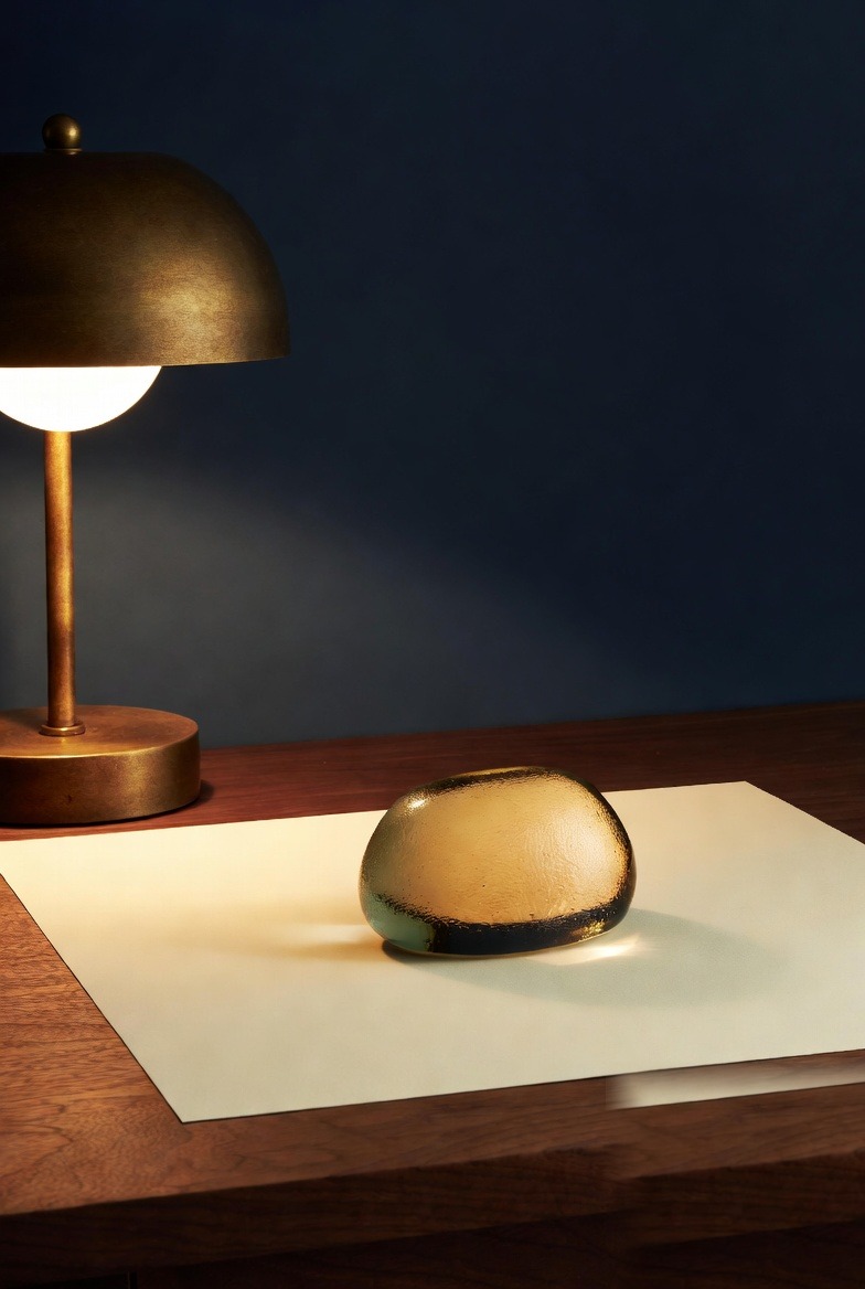A brass desk lamp and amber glass paperweight on a sheet of cream paper, set against a deep navy wall.